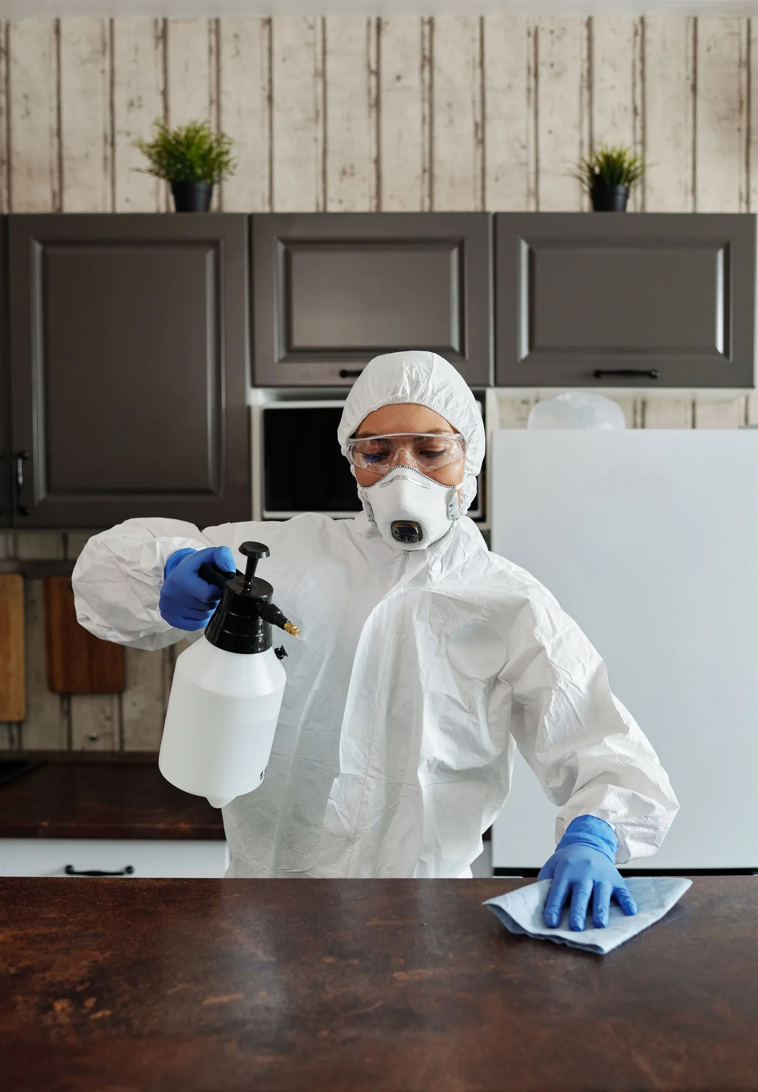A person in protective clothing disinfecting a table with a sprayer and cloth