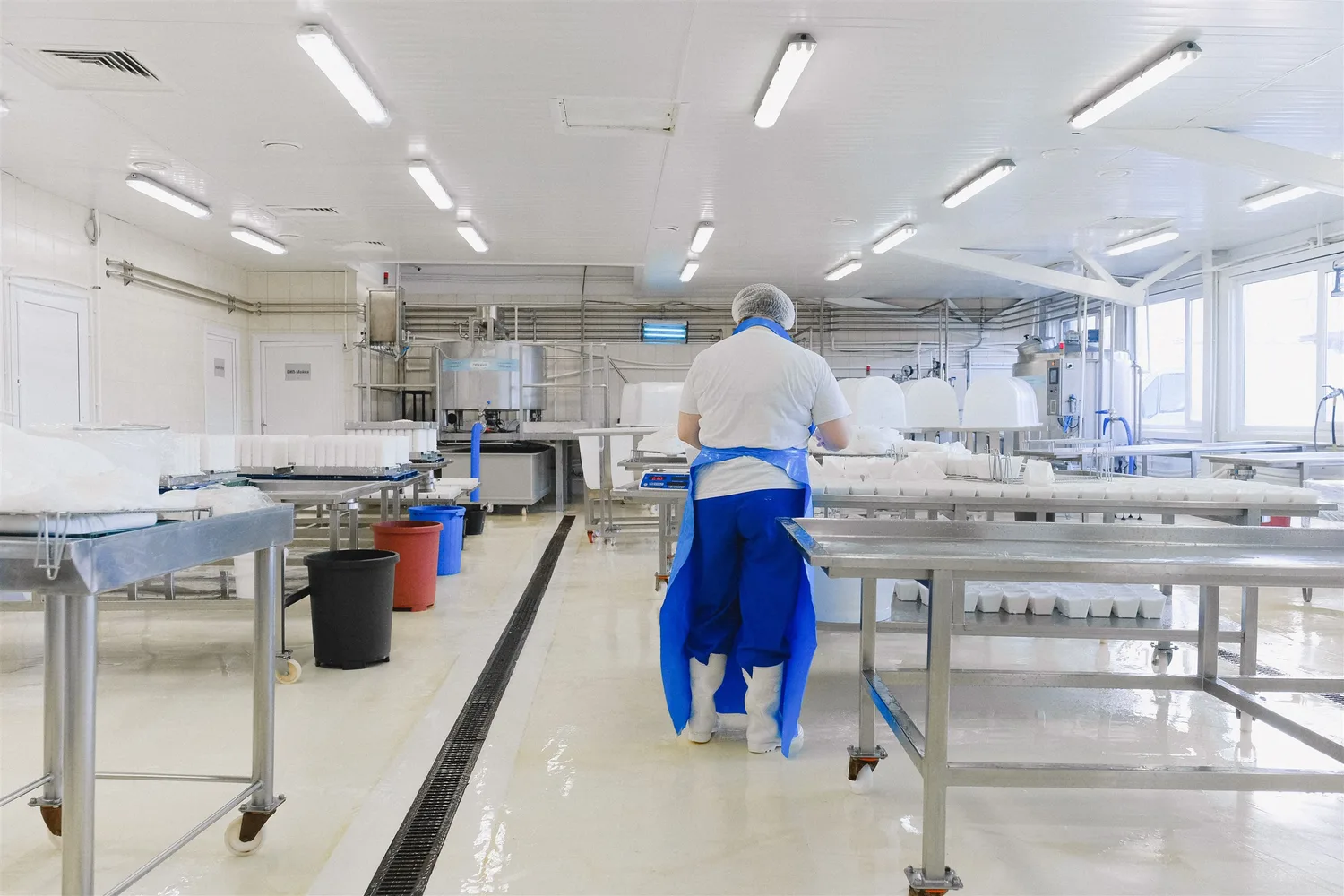 A food-production worker walking between stainless-steel preparation tables in a large kitchen
